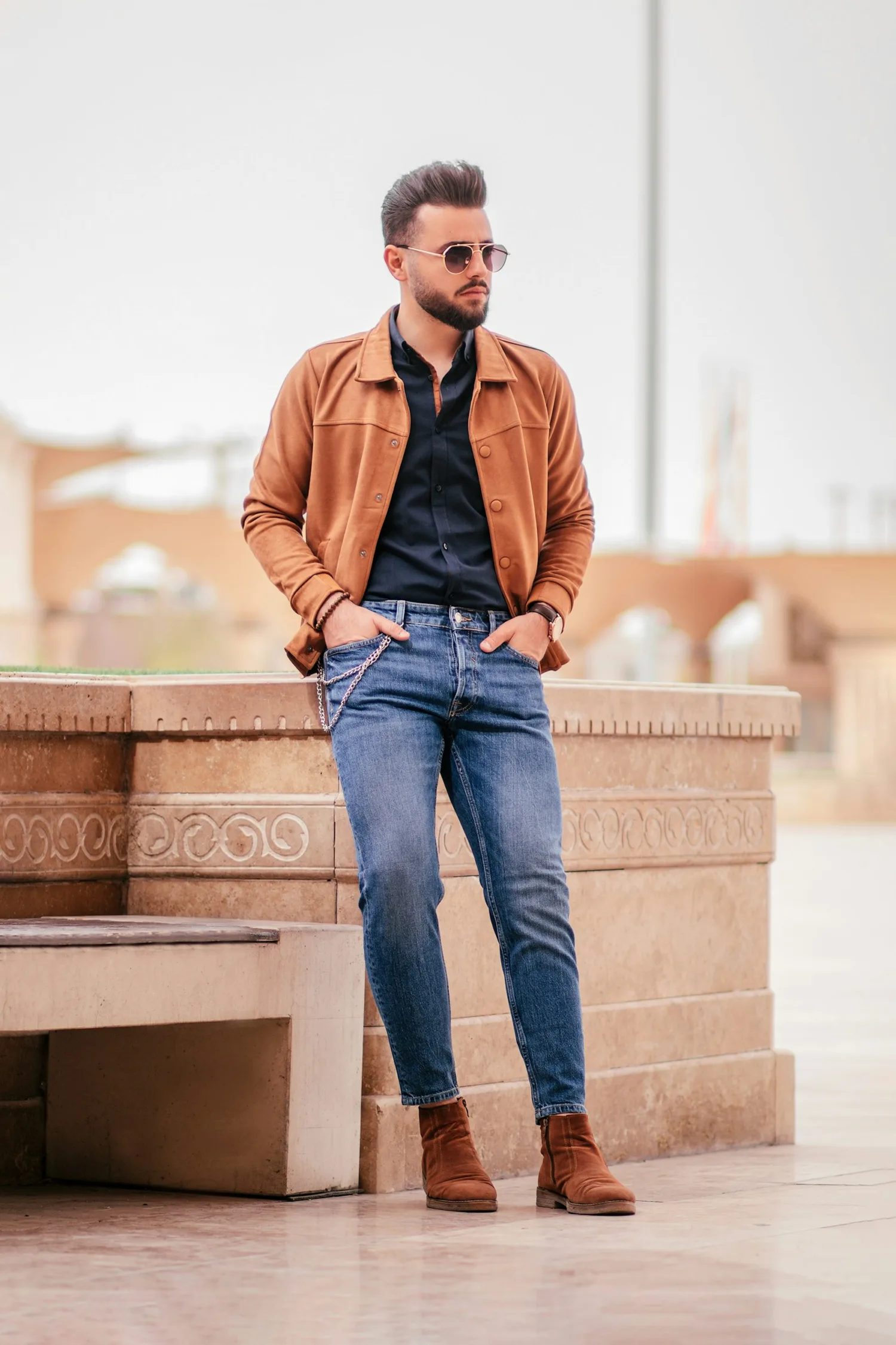 A man styling a tan jacket with a dark shirt, blue jeans, and brown boots